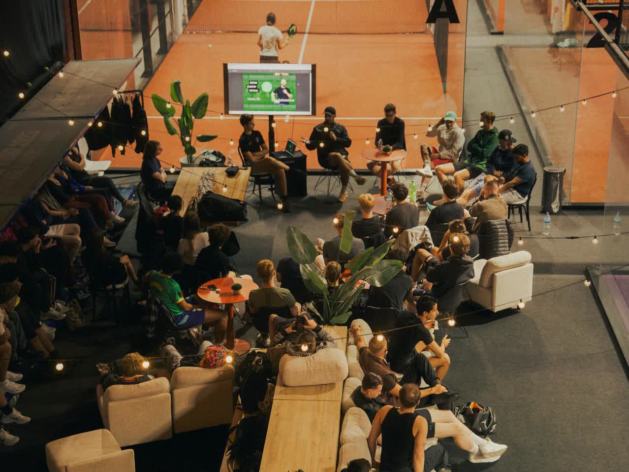 Abendveranstaltung in einer Padel-Halle: Ein Panel spricht vor Publikum auf Sofas, im Hintergrund ein beleuchteter Padel-Court